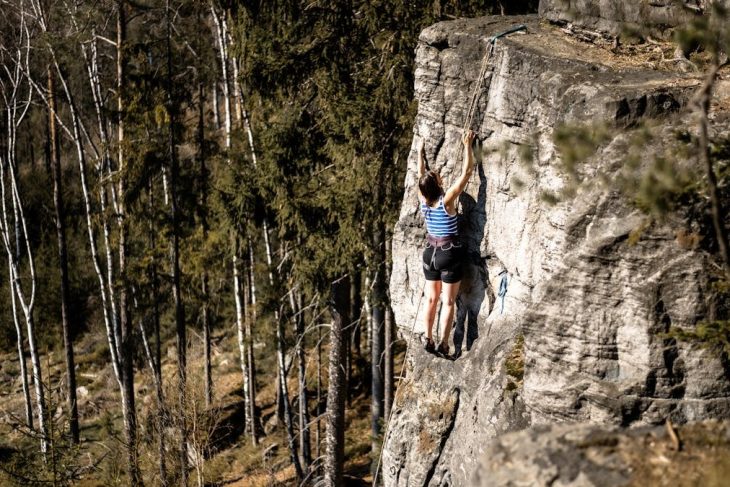 A woman ascending a rock face surrounded by a lush forest in Trutnov, Czechia.