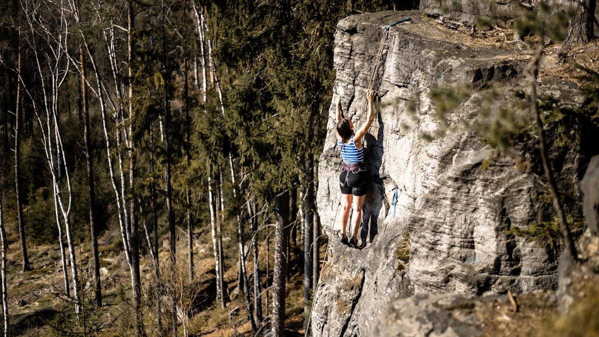 A woman ascending a rock face surrounded by a lush forest in Trutnov, Czechia.